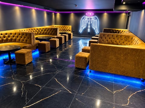 Modern lounge with gold tufted seating, neon wing sign, marble floors with LED lighting, and purple ambient lighting throughout the space