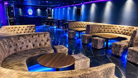 Modern nightclub lounge with gray upholstered seating, round wooden table, blue accent lighting, and polished dark floor